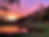 Scene with two-storey wooden house in midground surrounded by trees Sunrise on left with purple and rose pink and orange sky, green grass and autum leaves on trees.