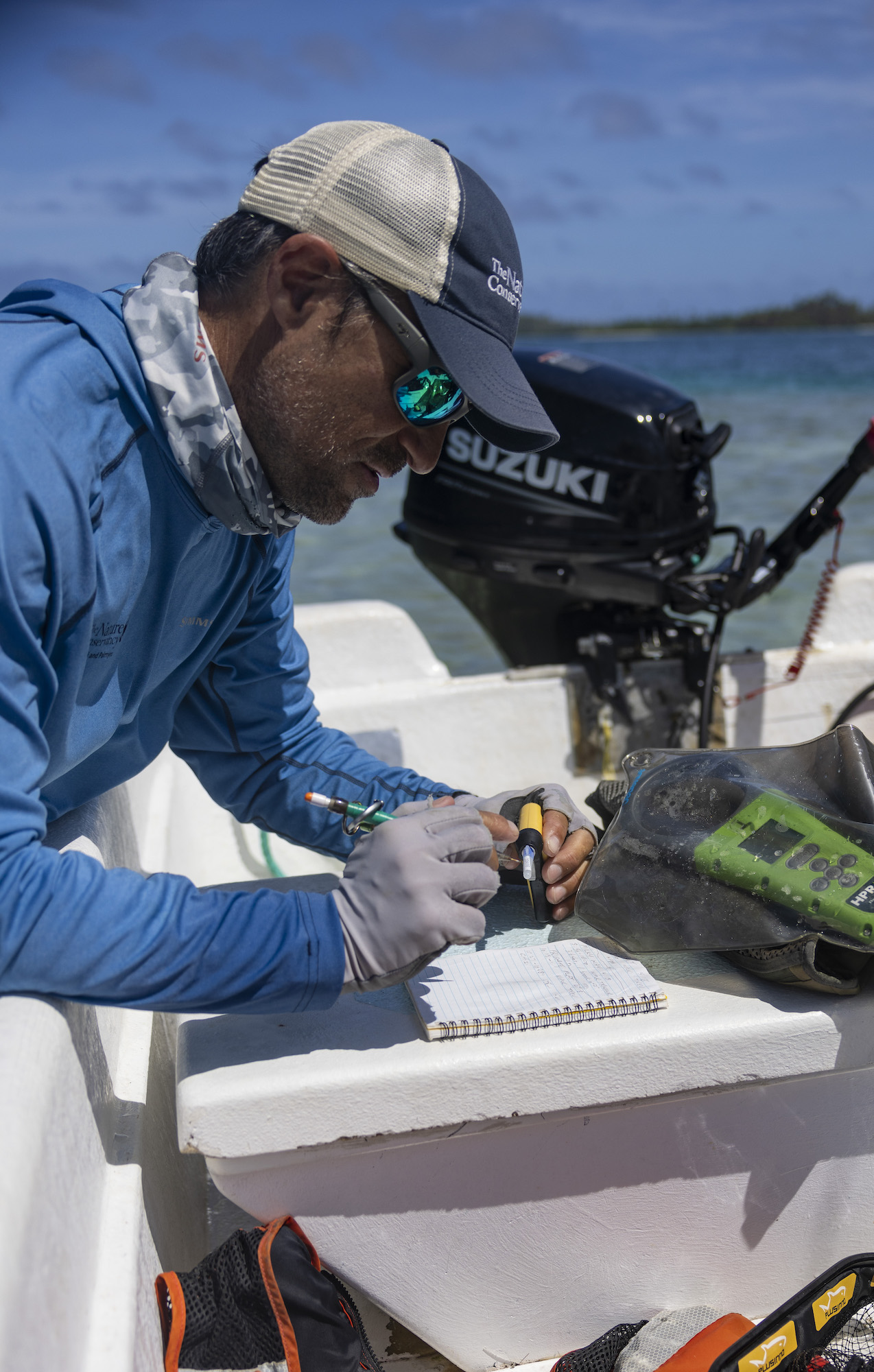 A man wearing fishing clothing, a hat and sunglasses takes notes on the back of a boat.