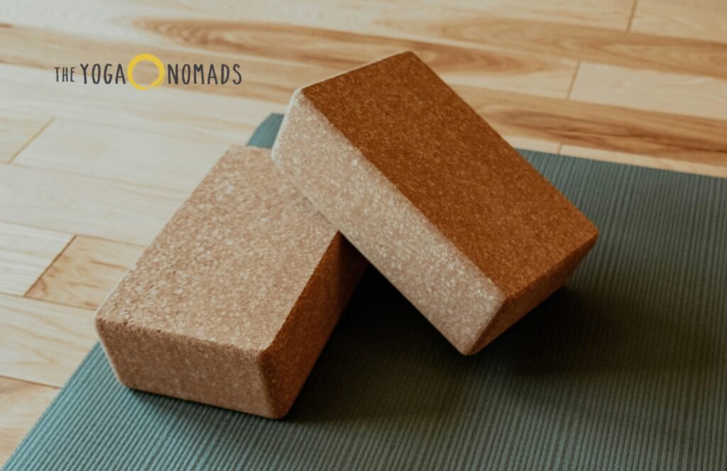 5 Essential Yoga Props for Joint Support During Practice yoga props for joint support 1 Two cork yoga blocks rest on a green yoga mat, arranged with one block leaning diagonally against the other to display their texture and form. The blocks sit on a wooden floor, and the text 'THE YOGA NOMADS' appears in the top left corner. This setup showcases how cork blocks offer sturdy, joint-supportive assistance during yoga practice by enhancing stability and alignment.