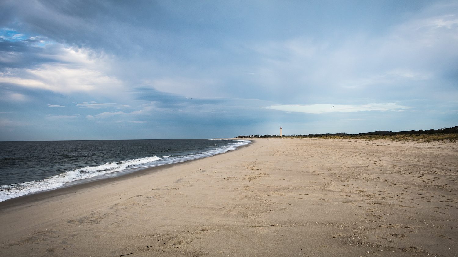 View looking down the beach at Cape May, with the ocean in the foreground and a lighthouse in the distance.