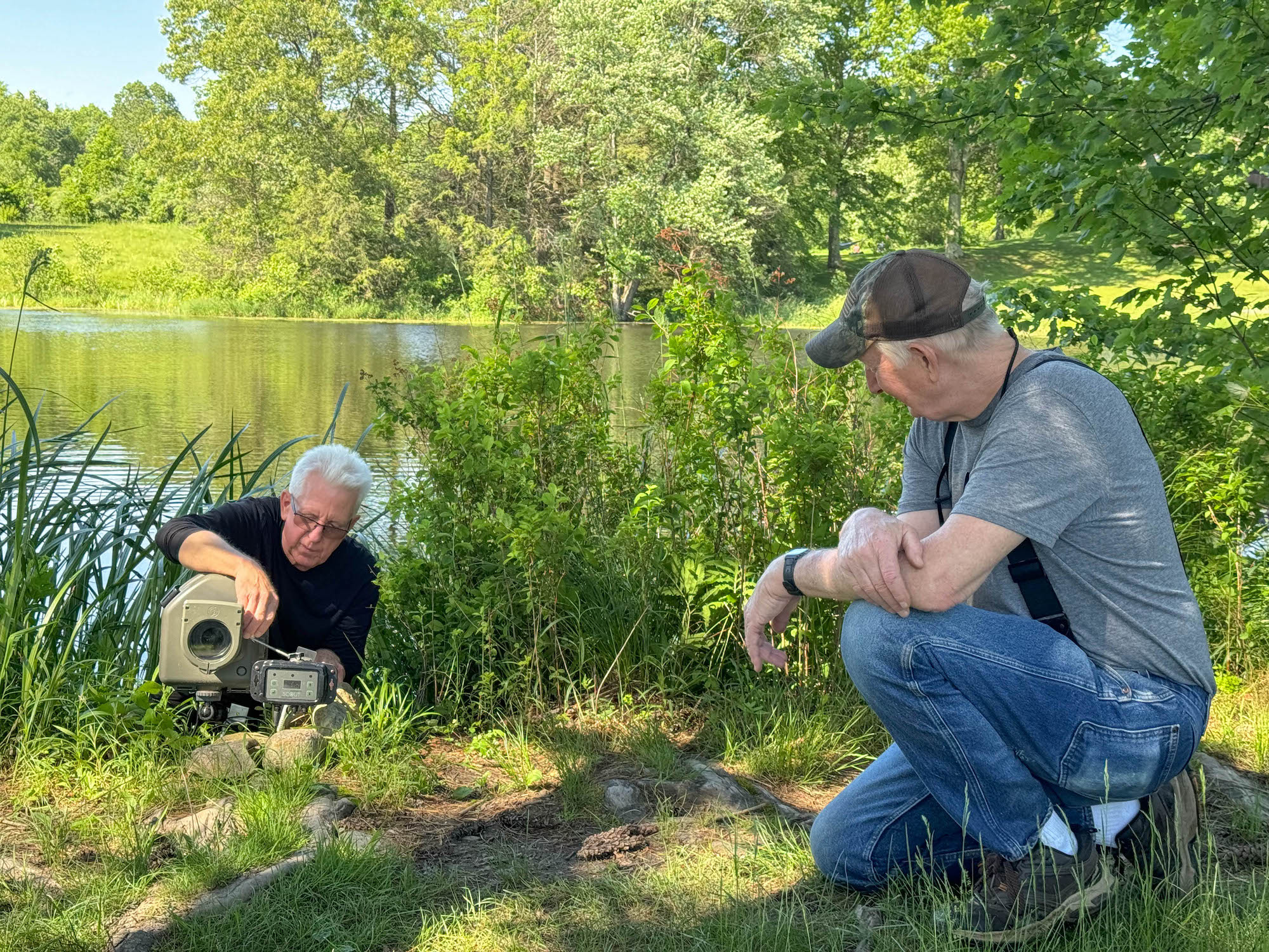 Two men set up a camera near a pond.