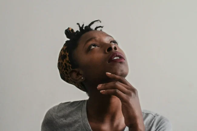 A black woman wearing a leopard print headscarf, under which mini braids are visible, has her hand on her chin and her head tilted back as she looks upward. A black woman wearing a leopard print headscarf, under which mini braids are visible, has her hand on her chin and her head tilted back as she looks upward.