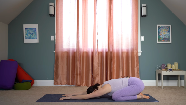 woman in purple yoga clothes in child's pose on yoga mat in home in front of window with closed curtains